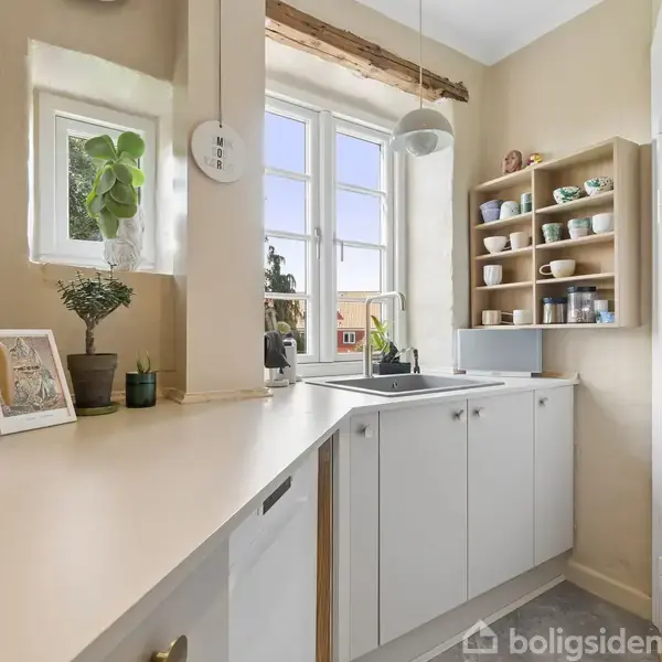 En stue med en sofa, et rundt spisebord og stole. Et stort vindue lader naturligt lys strømme ind. Væggene er dekoreret med malerier og en lampe står i hjørnet.