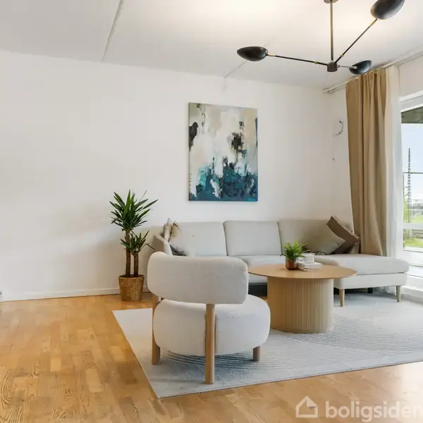 Sofa står i en lys stue ved siden af et rundt bord, omgivet af planter og kunst på væggen; store vinduer med gardiner åbner ud til en balkon. Ingen tekst fundet.