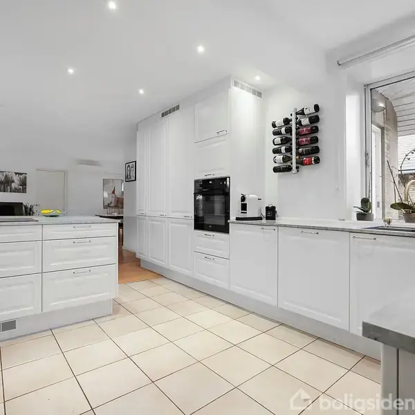 Et moderne køkken med hvide skabe og integreret ovn. Der hænger en vinreol på væggen. Store vinduer giver udsigt til en stenmur og himlen udenfor.