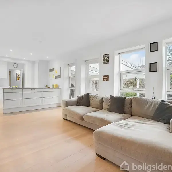 Sofaen står i en lys stue og vender mod det åbne køkken med ø. Store vinduer giver udsigt til en have, og lys strømmer ind, skaber en indbydende atmosfære.