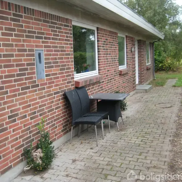 Et rækkehus af røde mursten med tre vinduer. To sorte stole står ved et lille bord på en flisebelagt sti langs huset. Grønt hegn og træer i baggrunden.