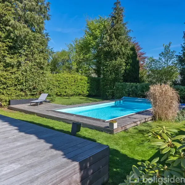 En swimmingpool omkranset af en træterrasse står stille i en velplejet have med høje, tætte træer og buske, der skaber en privat atmosfære. En solseng står ved poolens kant.