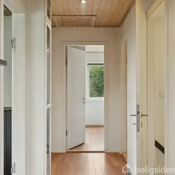 Gang med træloft og trægulv, døre åbne til flere rum. Væggen har teksten "Fremtiden er Fortiden". Naturligt lys strømmer ind fra et vindue i bagerste rum.