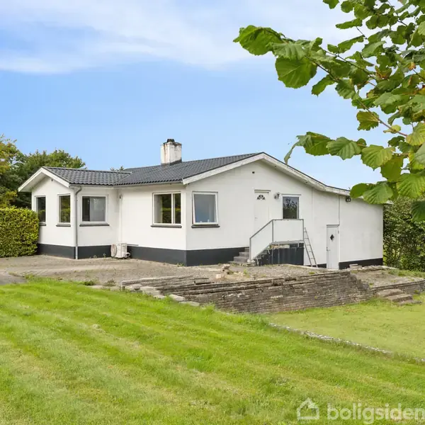 Et hvidt hus står stille på en grøn græsplæne med træer omkring. En stenbelagt trappe leder op til hovedindgangen, og himlen er blå i baggrunden.
