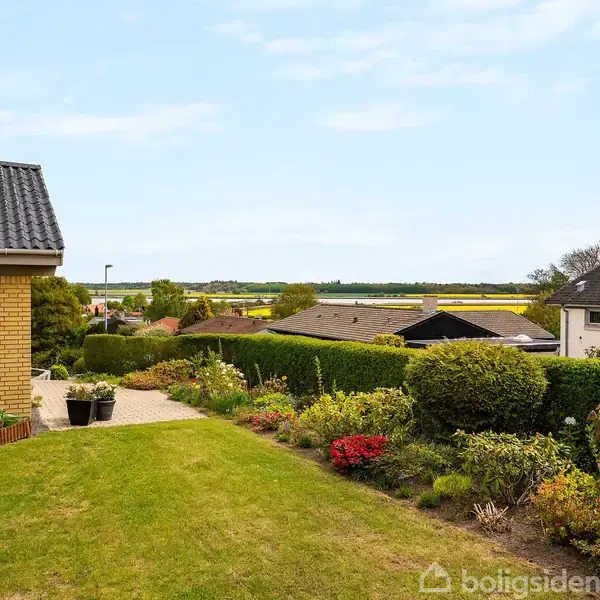 Et gult murstenshus med en græsplæne foran. Farverige blomster og buske dekorerer haven. Udsigten bagved viser flere huse og grønne marker i horisonten under en blå himmel.