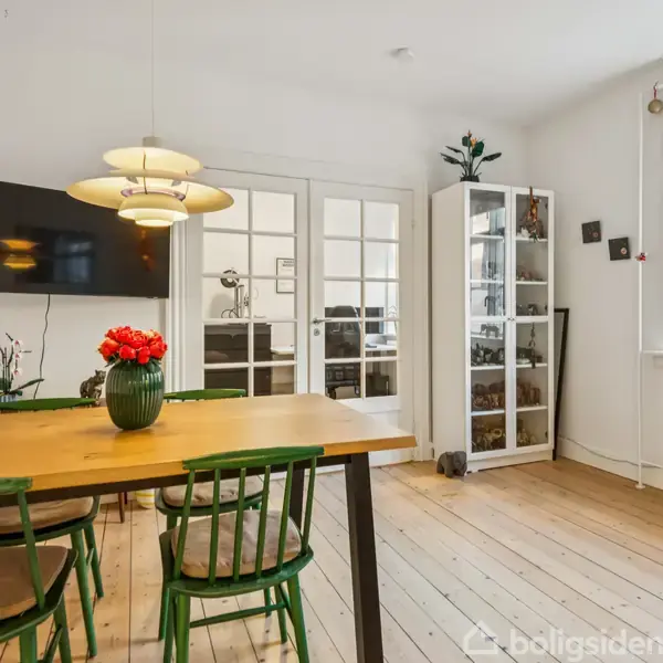 Et spisebord står i et lyst rum med grønne stole omkring sig. En vase med røde blomster er på bordet. Bagved er en glasvitrine og dobbeltdøre, mens et tv hænger på væggen.