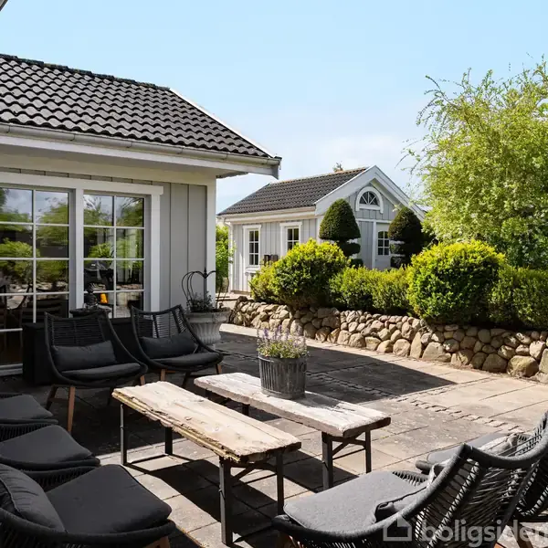 Terrassemøbler er arrangeret omkring et træbord på en flisebelagt terrasse. Baggrunden viser en træbeklædt bygning med store vinduer og omgivende grønne buske og træer.