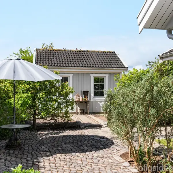 En lille, gråmalet bygning med hvide rammer står i en have. Foran er en brostensbelagt terrasse med en parasol og bord. Omgivet af buske og træer under blå himmel.