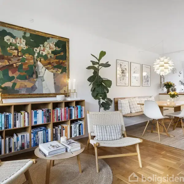 Stue med to stole og et rundt bord på trægulv. Væggen har et farverigt maleri, reol med bøger, og plante. Spisebord med stole i baggrunden nær et moderne køkken.