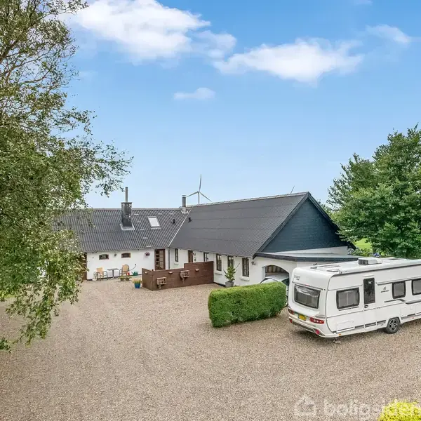En campingvogn holder parkeret på en grusplads foran et hvidt hus med sort tag. Omkring er der grønne træer og en blå himmel i baggrunden.