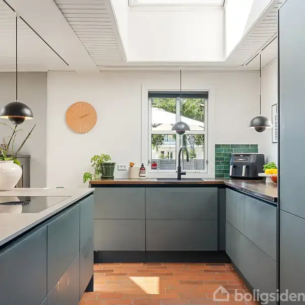 Køkkenet har blå skabe og en kogeø med hængende lamper. Et vindue i baggrunden, grønne fliser, planter og køkkenmaskiner fuldender det moderne og lyse design.
