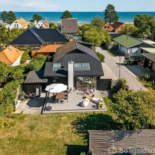 Hus beliggende i en grøn have med en terrasse og udsigt til havet i baggrunden. Terrasse indeholder parasol og siddepladser. Omkranset af andre huse og træer.