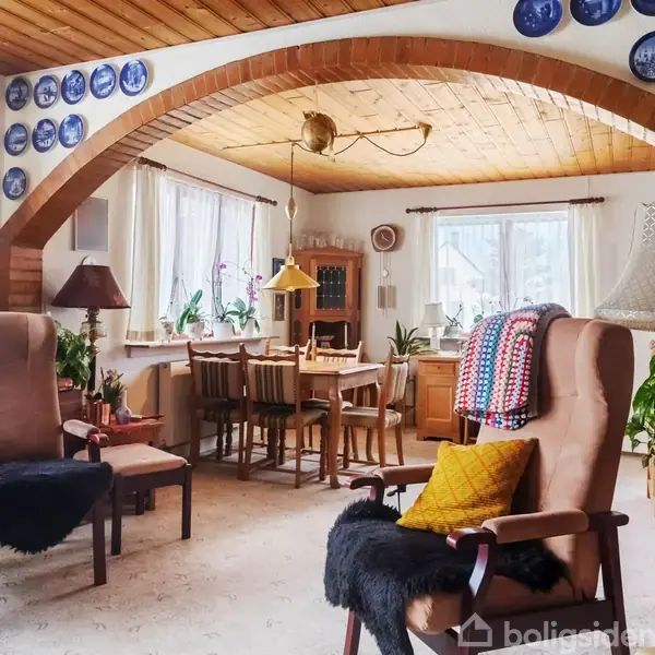 I et hyggeligt opholdsrum med træloft og -bjælker står to lænestole med puder og tæpper. Et spisebord med stole er opstillet nær vinduer, omgivet af planter og dekorative tallerkener.