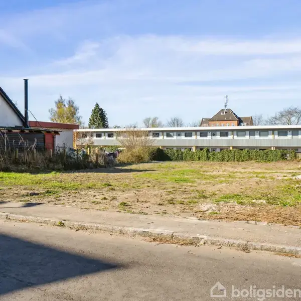 Tom grund står ubenyttet midt imellem to huse. En husrække er synlig i baggrunden, og en informationsskilt er placeret tæt på grunden. Himmel er klar og solrig.
