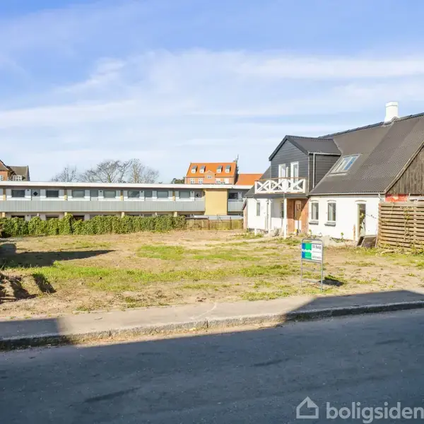 Et sort og hvidt hus står stille ved siden af en tom grund, omgivet af lave hegn og en asfaltvej. I baggrunden ses andre bygninger og en blå himmel.