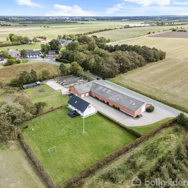 Hus med rødt tag og tilhørende staldbygninger ligger ved vejene, omgivet af grønne marker og skov. Flere huse kan ses bagved. Grønt område med hæk omkranser ejendommen.