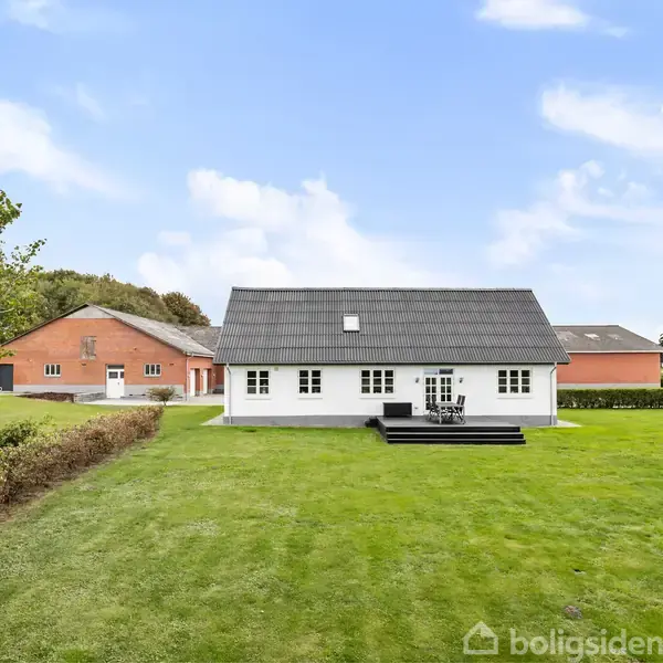Et hvidt hus med mørkt tag står i en grøn have. Bagved er en rød udbygning. Himlen er klar med få skyer.