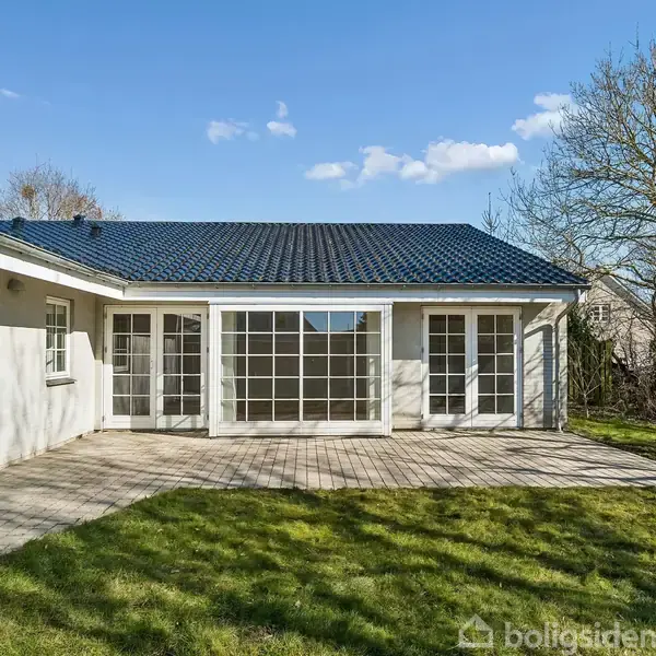 Et hvidt hus med store vinduespartier og sort tag står stille på en flisebelagt terrasse. Omkring huset ses en grøn græsplæne og bare træer under en blå himmel.