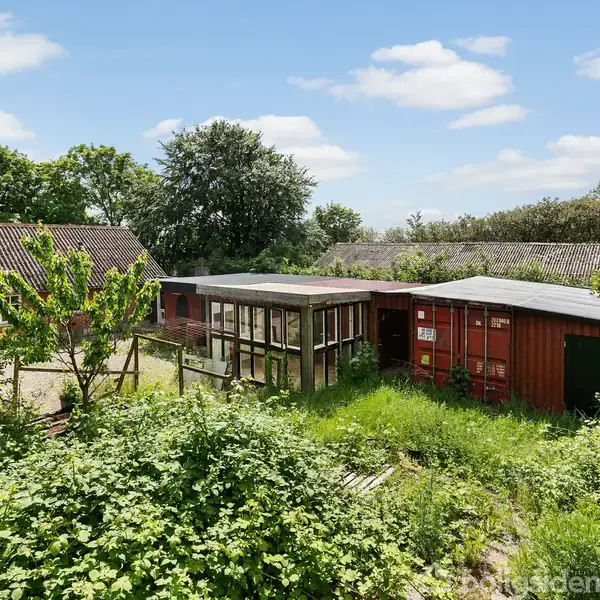 Rødt træhus står i en frodig have med masser af grøn vegetation. Omkring huset er der flere træer og en blå himmel med spredte skyer.