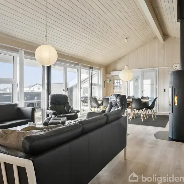 Sort lædermøbelarrangement står stilrent i stue, mens brændeovn afgiver varme. Store vinduer lukker lys ind over moderne spisebord. Træloft tilføjer rustik charme. Korridor fører til andre rum.