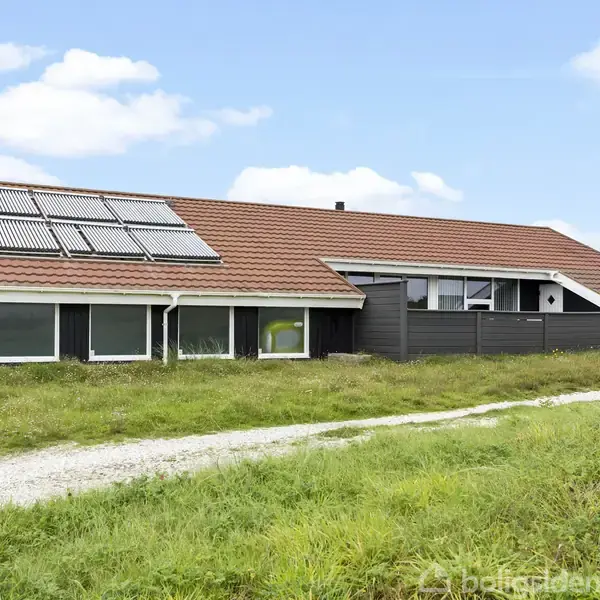 Et rækkehus med rødt tegltag og solpaneler står i et åbent grønt område. En grussti fører op til huset, omgivet af græs og enkelte buske.