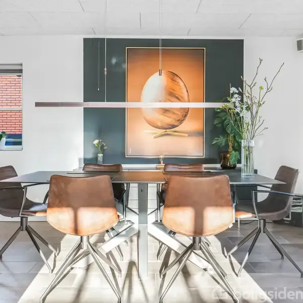 Spisebord med seks læderstole i et lyst køkken/alrum. Lang lampe over bordet. Væggen har et stort billede af et æg. Værelset har vinduer med planter i vindueskarmen.