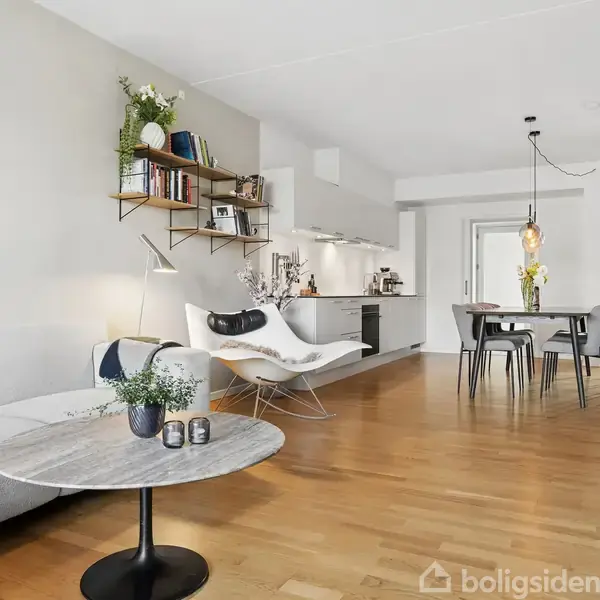 En moderne stue med en lys sofa i venstre side, der står foran et rundt marmorbord. Til højre er et spisebord med stole. I baggrunden ses et åbent køkken.