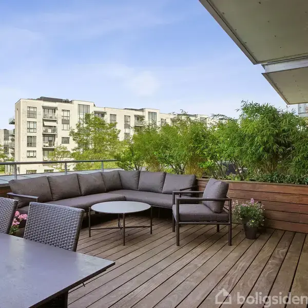 Terrasse med hjørnesofa og bord. Komfort er omdrejningspunktet. Rundt omkring er der træ og buske som baggrund. Flere etagebygninger ses i baggrunden under en blå himmel.