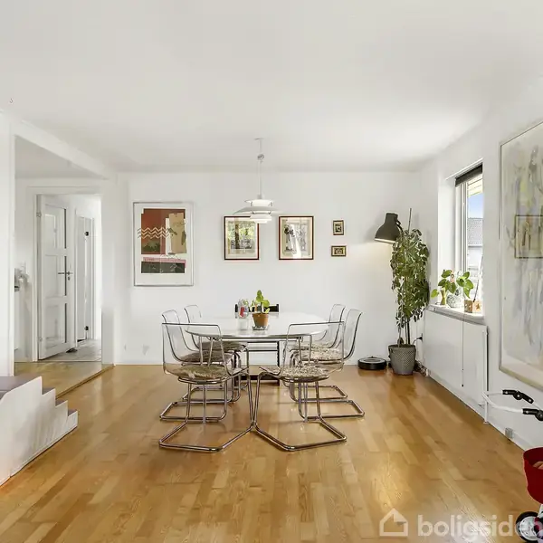 Et moderne spisebord med seks gennemsigtige, armløse stole står i et lyst rum. På væggene hænger flere billeder. En stor plante står ved siden af vinduet, som slipper sollys ind.