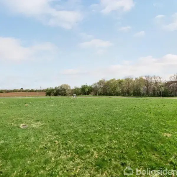 En stor, grøn græsplæne breder sig ud mod en række træer i baggrunden. En asfaltsti løber langs græsset i et åbent naturområde under en skyet himmel.