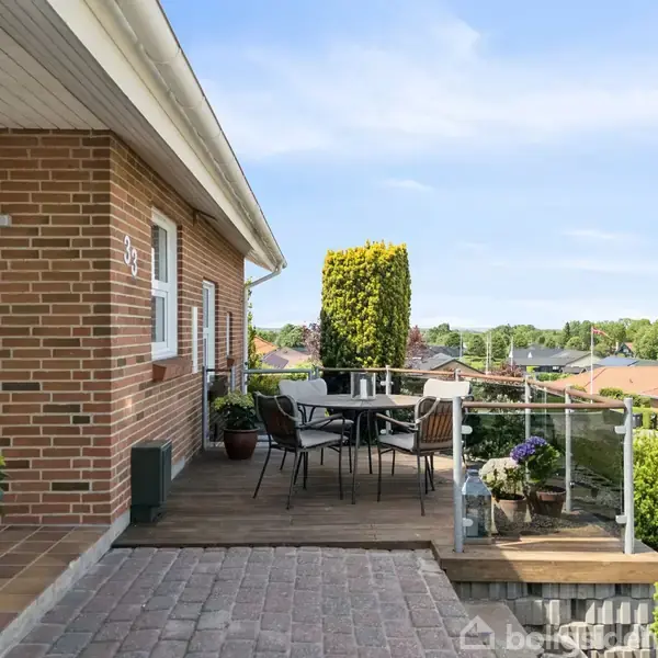 Murstenhus med terrasse og glasgelænder. Terrassebord med stole står på trædæk. Planter i krukker skaber en hyggelig atmosfære. Udsigt over parcelhuskvarter og træer i baggrunden. Husnummer 37.