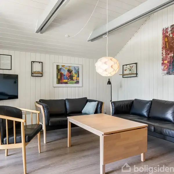 Et moderne stueområde med sorte lædersofaer og træstole omkring et træsofabord. En fladskærm er vægmonteret, og kunst dekorerer væggene. En lampe hænger fra loftet.