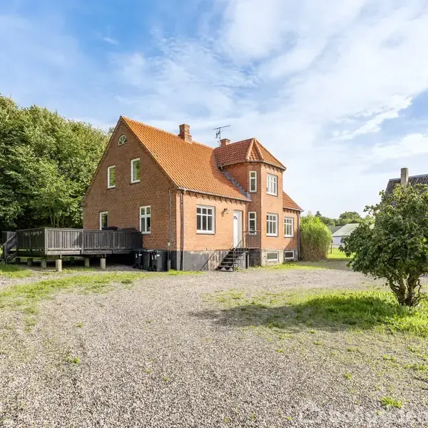 En rød murstensvilla står stille i en stor grusbelagt gårdsplads omgivet af grønne træer og en græsplæne under en blå himmel.