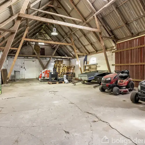 Traktorer holder stille i en rummelig, åben lade med højt til loftet af træ og metal. Gulvet er beton, og omgivelserne inkluderer paller og forskellige landbrugsudstyr i baggrunden.