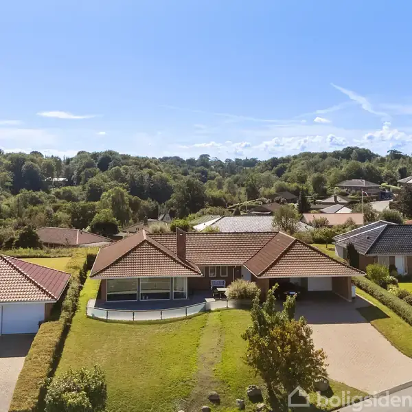 Huset står stille på en række parcelhuse omgivet af velholdte haver og grønne træer, i et naturskønt boligområde under en klar blå himmel.