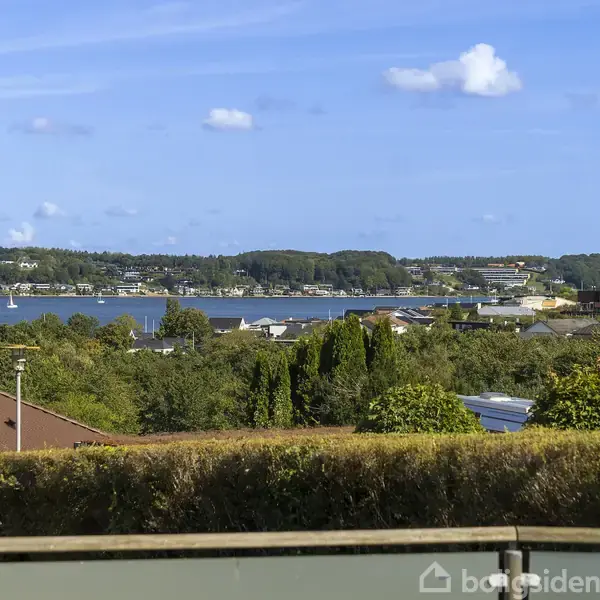 Havudsigt over en kystby, med flere bygninger og grønne områder i forgrunden, under en blå himmel med hvide skyer.
