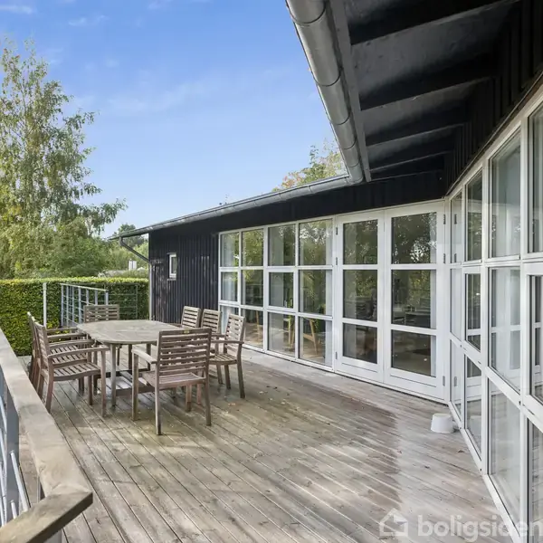 Terrassebord og stole står stille på en træterrasse ved siden af et mørkt hus med store glasvinduer, omgivet af grønne træer og buske under en klar himmel.