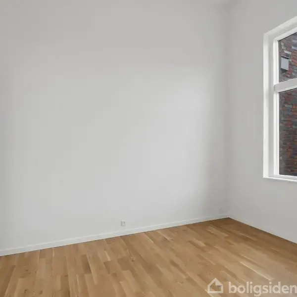 Et tomt rum har et hvidt skab og trægulv, mens store vinduer slipper lys ind, med udsigt over en bygningsfacade og bybillede udenfor.