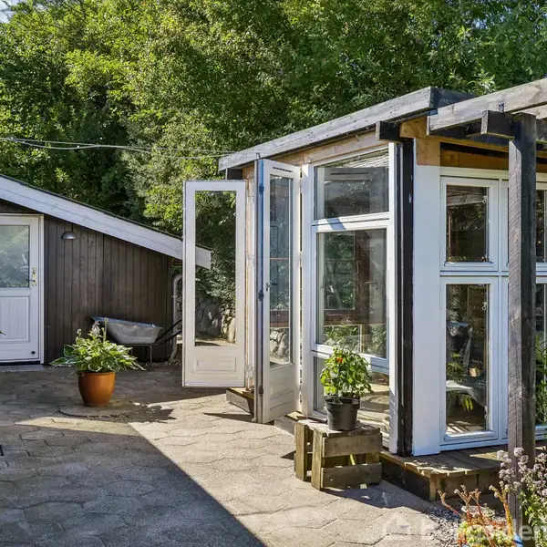 Et lyst orangeri med glasvægge står åbent, omgivet af en flisebelagt terrasse. I venstre side ses en mørk bygning med en hvid dør. Grønne planter fylder omgivelserne.