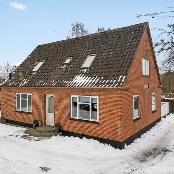 Rødstenshus med mørkt tag ligger i et snedækket landskab. Huset har flere vinduer og en hoveddør med små buske ved siden af. Et kirketårn ses i baggrunden.