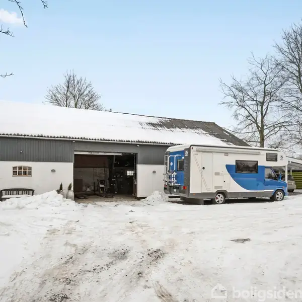 En kampvogn står parkeret foran en stor, sneklædt lade med åben port. Træer uden blade omkranser området, og himlen er lys og klar.