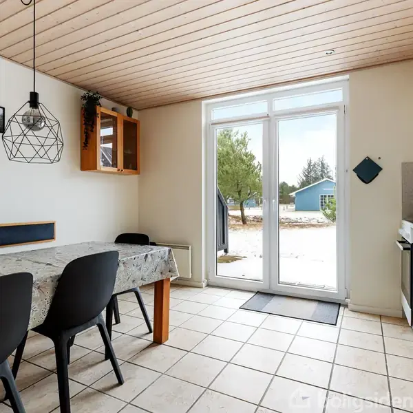 Et moderne køkken-alrum med et spisebord dækket med en dug, to sorte stole og en stor glasdør, der fører ud til en gårdhave med træer i baggrunden.