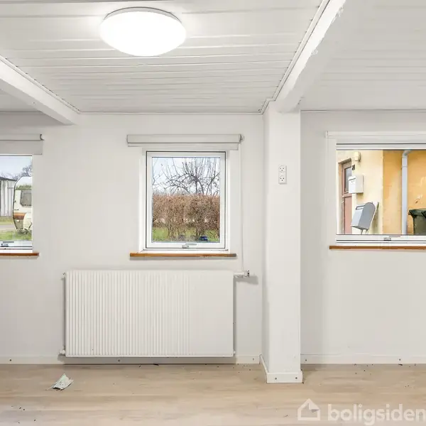 Værelse med hvidmalede vægge og loft samt tre vinduer, hvoraf to viser en have og et et gult hus. Der er et radiator under midtervinduet og trægulv.