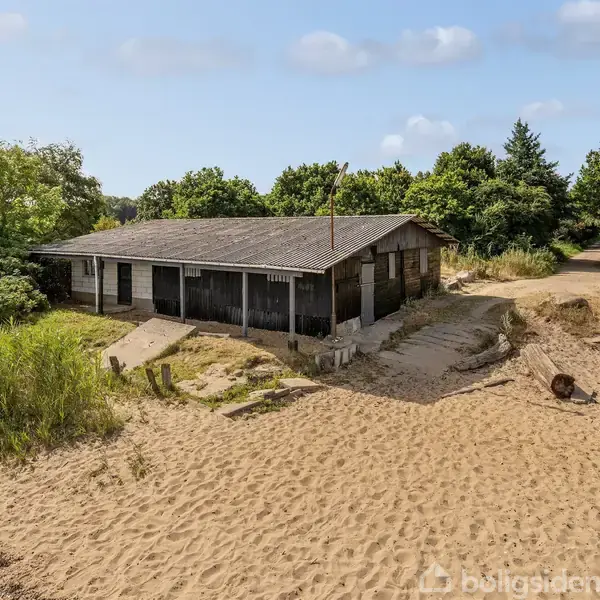 Et gammelt, træhus står stille omgivet af sand og spredte buske. I baggrunden ses en frodig, grøn skov, og himlen er let skyet.