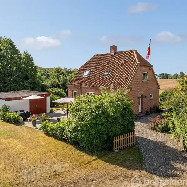 Murstenshus med rødtag står i sollys med en dansk flagstang. En have omgiver huset, og en grussti leder til indgangen. En carport ses til venstre.