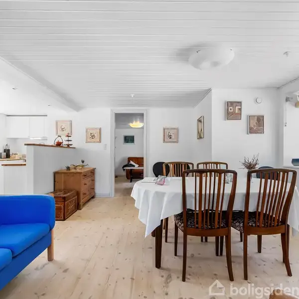 Et lyst rum med et træbord dækket af en dug, omgivet af seks stole. En blå sofa står ved vinduet. Væggene er dekoreret med billeder.