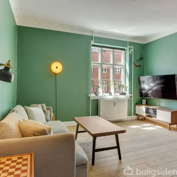 Stue med grå sofa, træbord og vægmonteret fjernsyn. Grønne vægge og stort vindue lader lys ind. Hyggelig atmosfære med enkelte dekorationer og radiator under vindue.