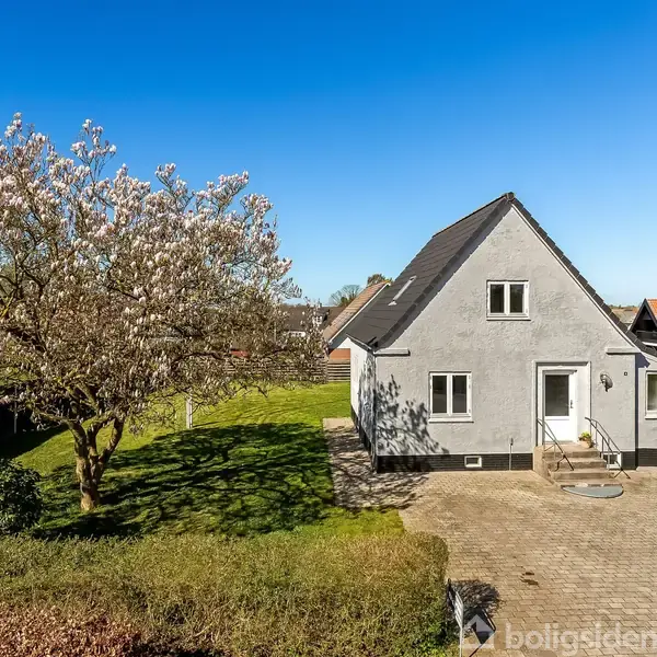 Et gråt hus med en enkeltdør og skråtag står i en have med et stort blomstrende træ. Belægningen foran huset danner en parkeringsplads.