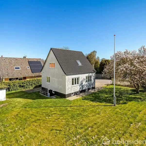 Et en-familiehus står på en stor, velplejet græsplæne med en flagstang og blomstrende træer i forhaven. Nabohuse og en klar blå himmel danner den omkringliggende kontekst.