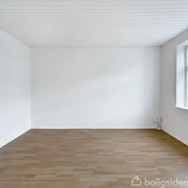 Et tomt rum med et trælaminatgulv og hvide vægge. Et vindue til højre lader naturligt lys komme ind. En radiator er placeret på venstre væg.